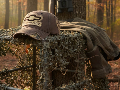 T-Top Vibes khaki Chevrolet cap with embroidered logo and frayed camo patch resting on a hunting blind in the woods.